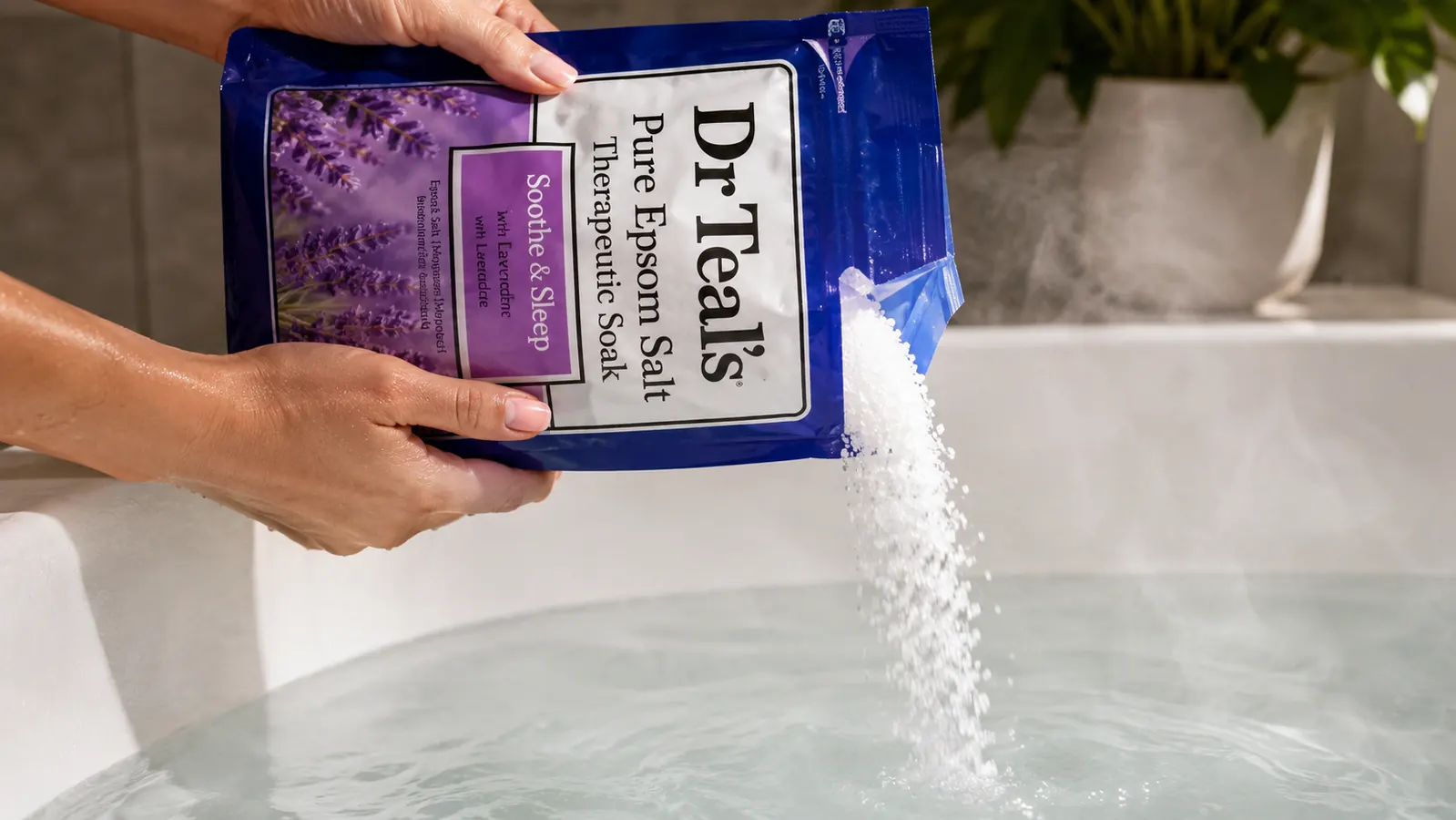 Two hands pouring epsom salt crystals into a hot bath, crystals dissolving in the water